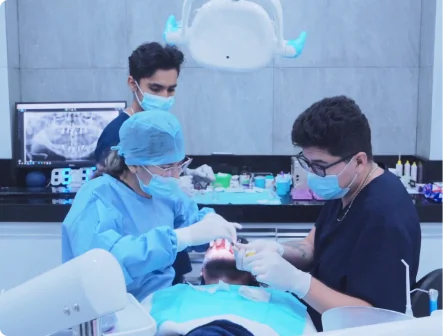 A team of three dental professionals wearing scrubs and face masks performing a dental implant procedure on a patient, with a dental x-ray visible on a monitor in the background.
