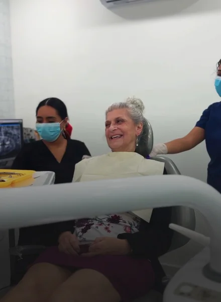 Happy senior female patient smiling in a dental chair with a clinic assistant in the background.